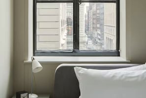 Et hotelværelse med en seng, et natbord, en lampe og et vindue med udsigt til byens bygninger.
