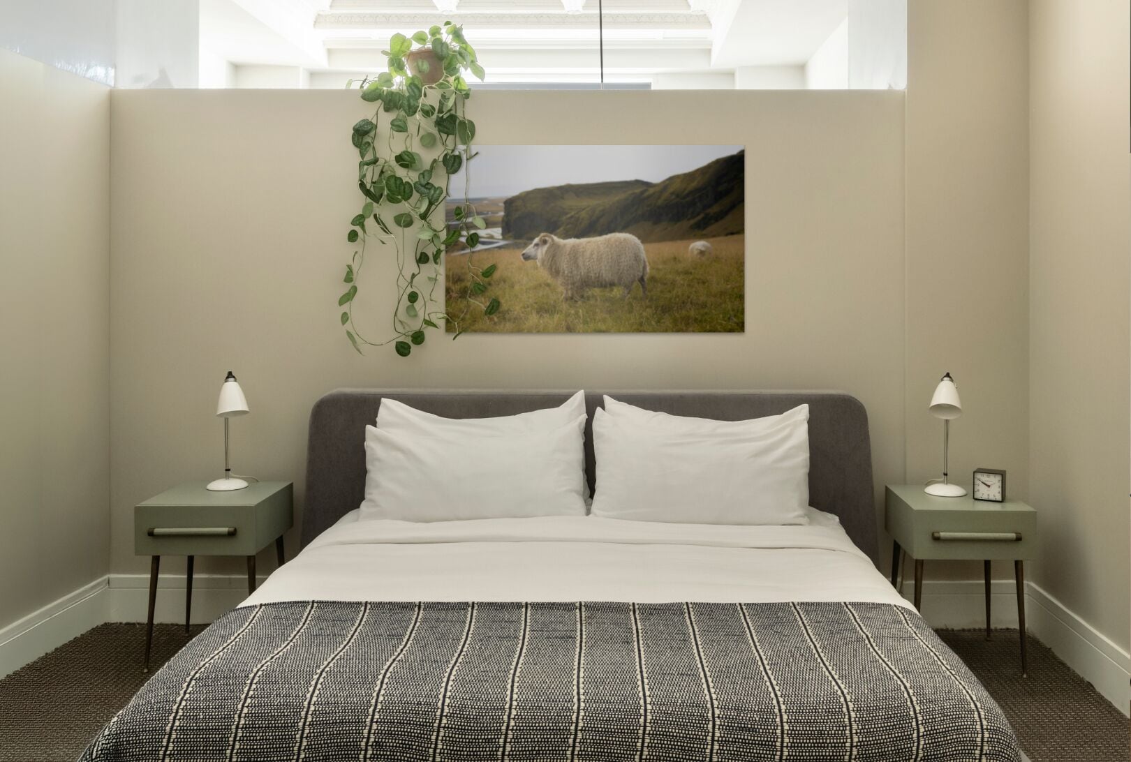 A bedroom with a large bed, two bedside tables with lamps, a wall-mounted plant, and a painting of sheep in a field.