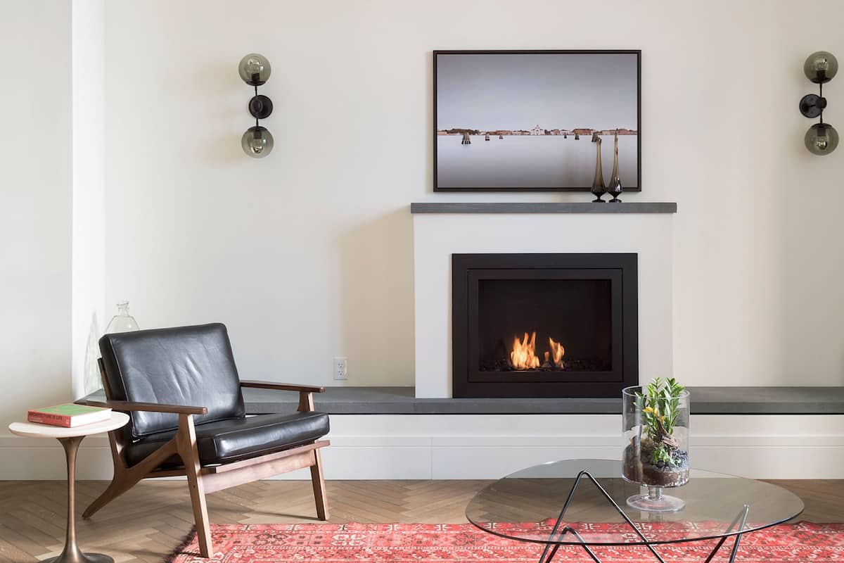 A living room with a fireplace, a black leather chair, a glass coffee table, and a red patterned rug.