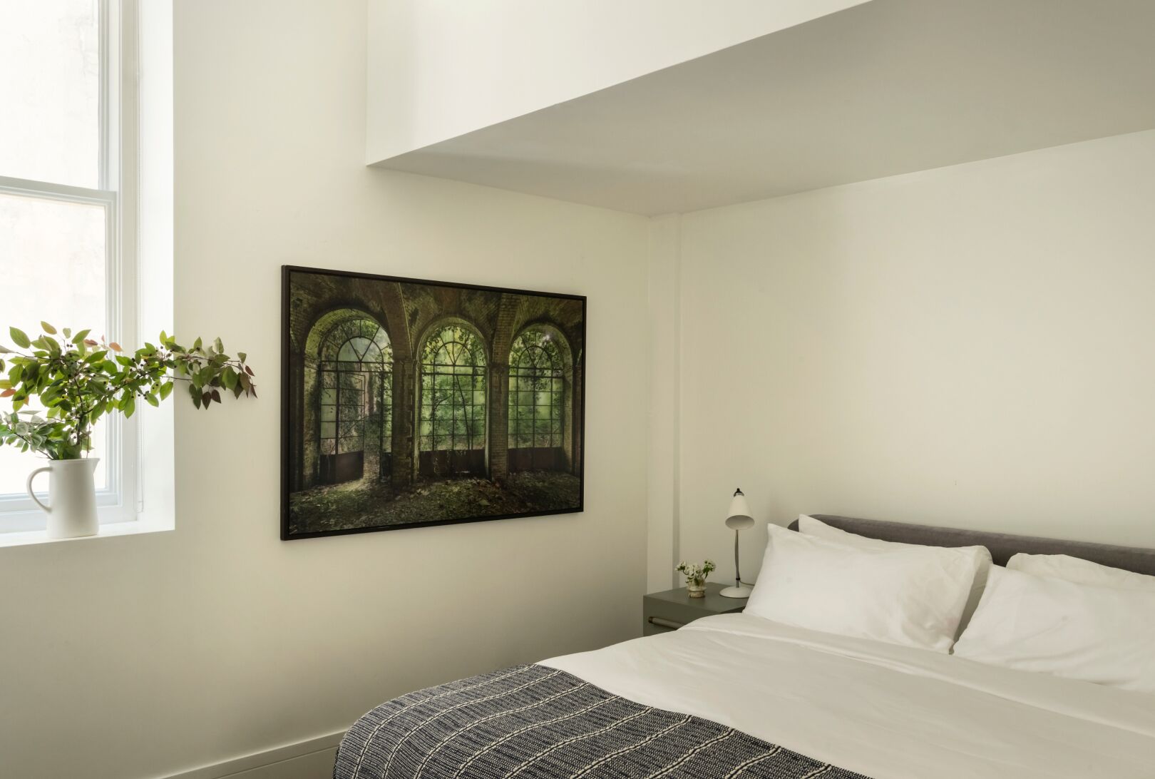 A bedroom with a bed, a nightstand, a lamp, a window with a plant, and a painting of a forested archway.
