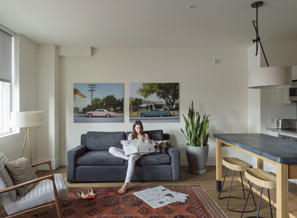 A modern living room with a grey sofa, a coffee table, a dining area with bar stools, and a kitchen in the background. - ROOST Midtown (Philadelphia)