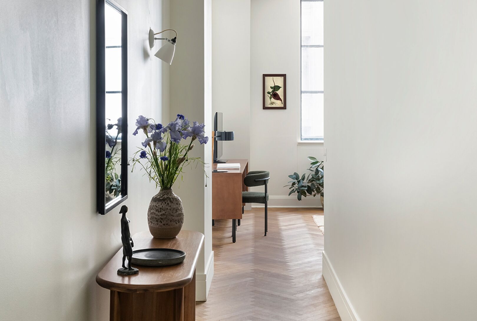 Un couloir avec un plancher en bois, un vase rempli de fleurs, un tableau encadré et un bureau avec une chaise.