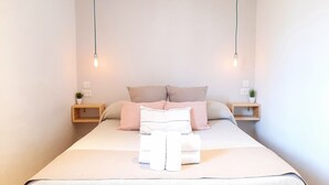 Een opgemaakt bed met kussens en een gevouwen handdoek, twee nachtkastjes met kleine plantjes, en twee hangende lampen.