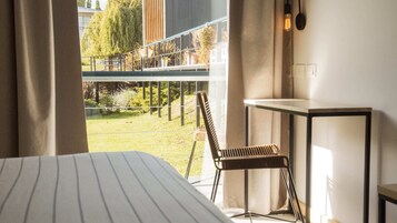 Habitación de hotel con una cama, una silla, una mesita y vistas a un balcón con plantas y un edificio al fondo.