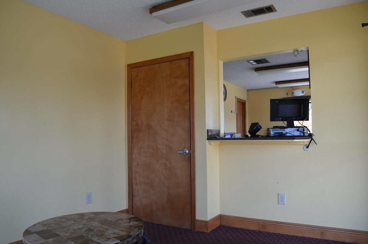A room with a wooden door, a small round table, a built-in shelf with a computer setup, and a wall-mounted mirror.