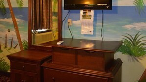 A room with a wooden dresser, a flat-screen TV mounted on the wall, and a mural of a beach scene. - Economy Inn (New Orleans)