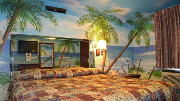 A bedroom with a beach-themed mural, a bed with a colorful quilt, a nightstand, and a lamp. - Economy Inn (New Orleans)