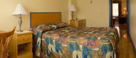 A hotel room with a wooden bed, two bedside tables with lamps, a wooden chair, and a visible door leading to another room.