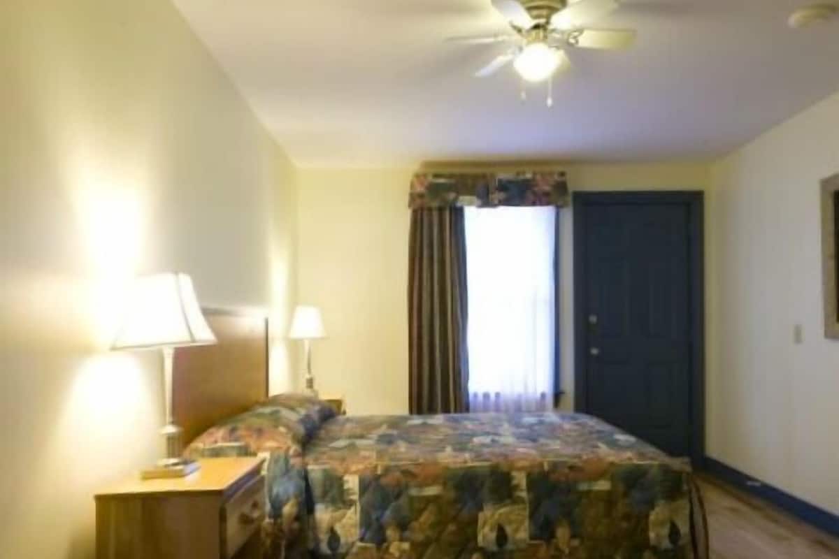 A hotel room with a bed, two bedside lamps, a ceiling fan, a window with curtains, and a door.