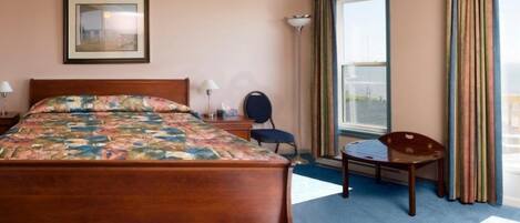 A hotel room with a wooden bed, a desk, a chair, a large window with curtains, and a framed picture on the wall.