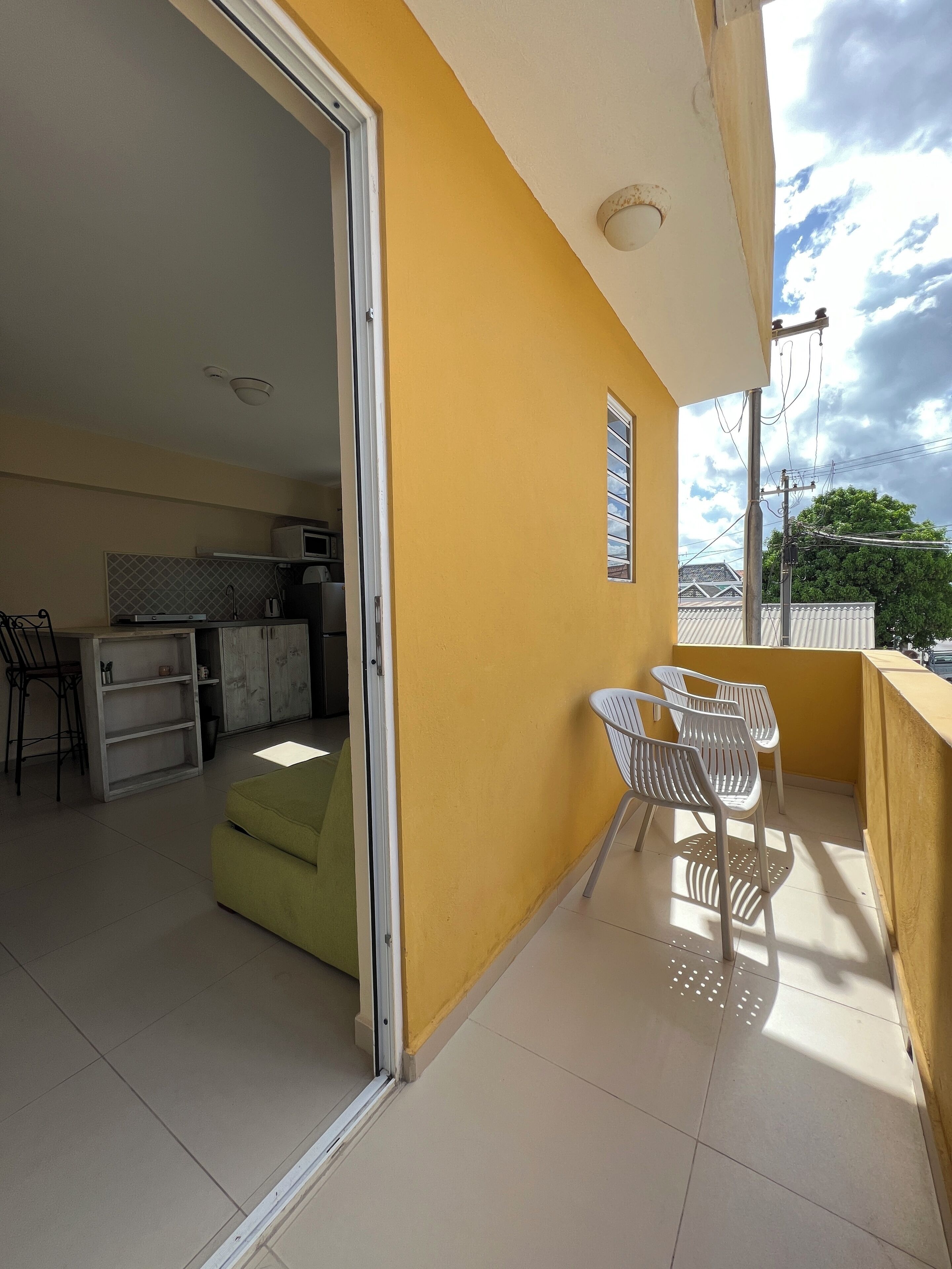 Un balcón con una pared amarilla, un ventanal corredera que da a una cocina y un pequeño comedor con sillas.