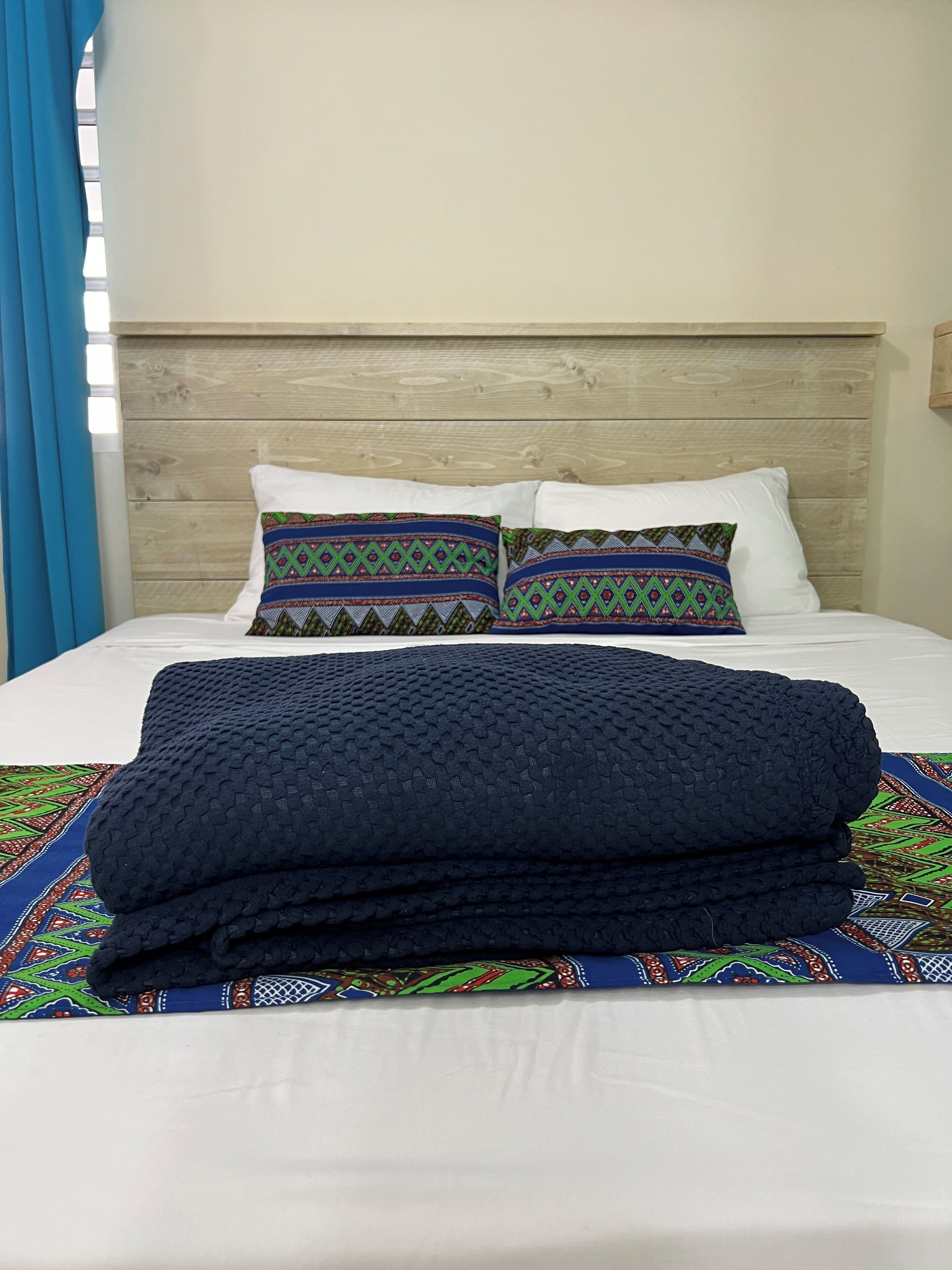 Una cama con cabecera de madera, dos almohadas estampadas, una manta doblada y una colcha colorida.