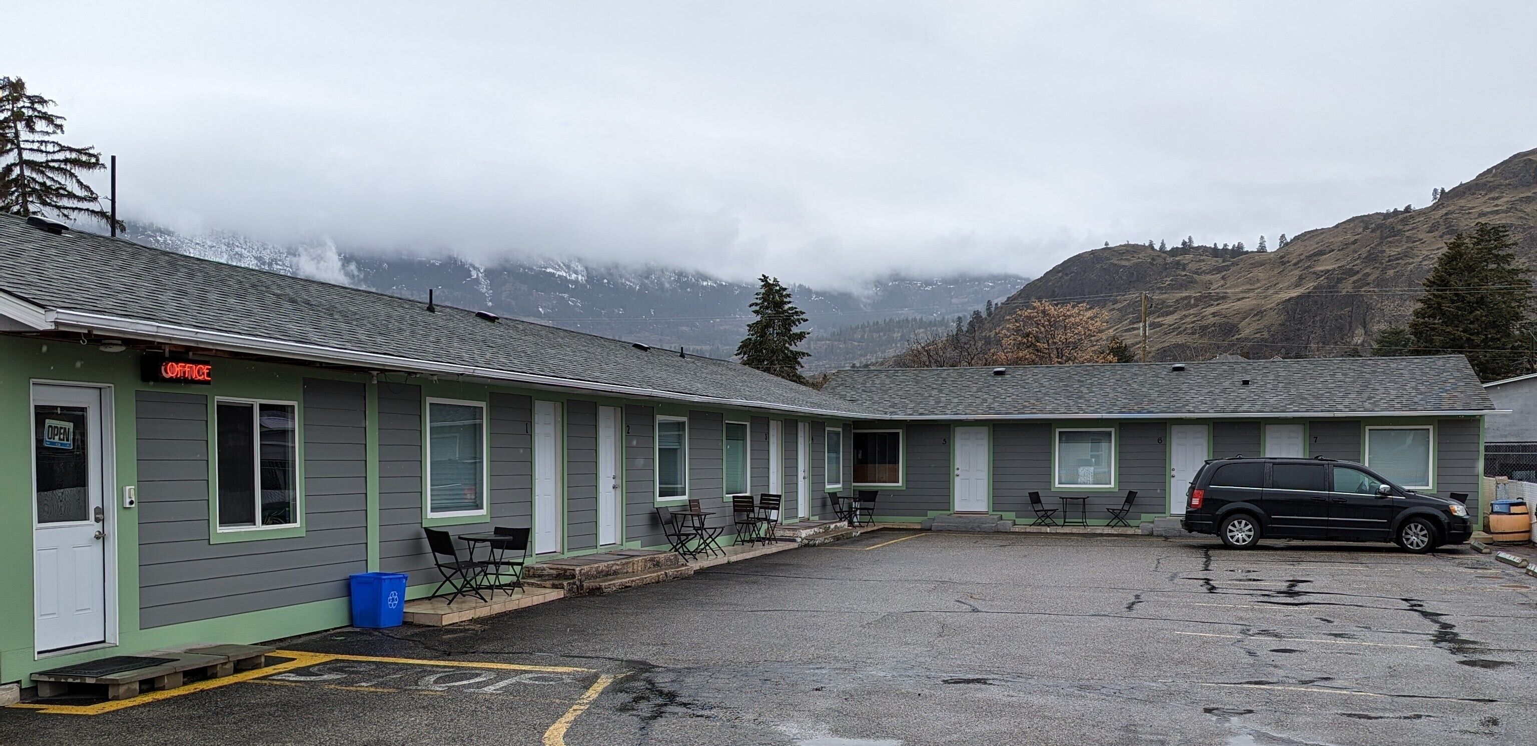 Un motel con fachada verde, varias habitaciones, asientos exteriores y un área de estacionamiento.