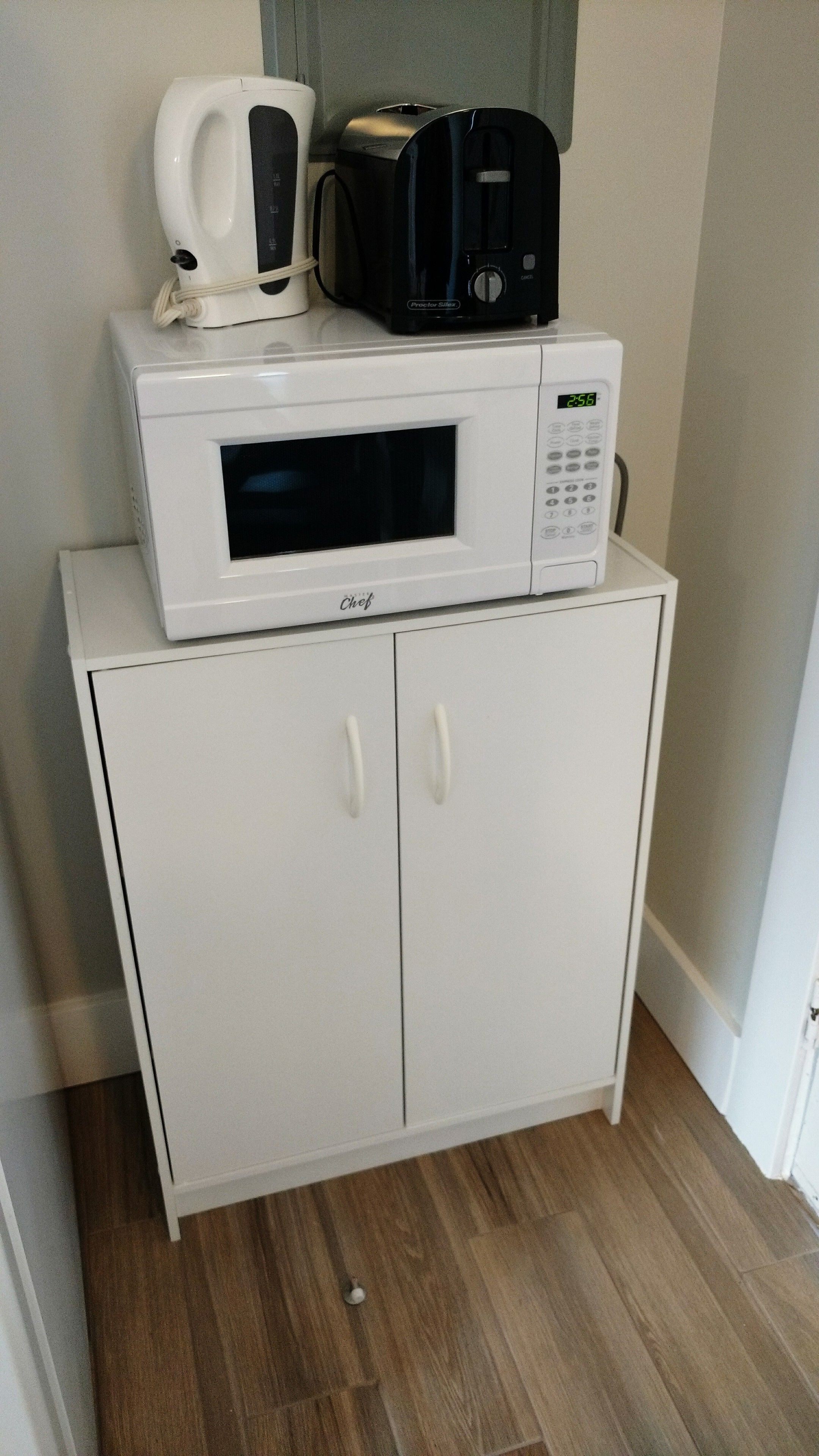 A white microwave oven with a digital display and a coffee maker on top, placed on a white cabinet with two doors.