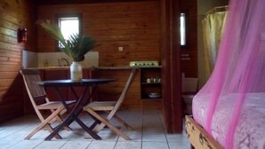A compact cabin-style room with a wooden interior, a dining table with chairs, a small kitchenette, a single bed with a pink canopy, and a window with a view. - 3 Rivers & Rosalie Forest Eco Lodge (Rosalie)