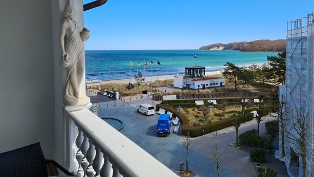 A balcony with a view of the sea, a white building with a blue van, and a statue. - Aparthotel Rialto & Hotel Esplanade (Binz)
