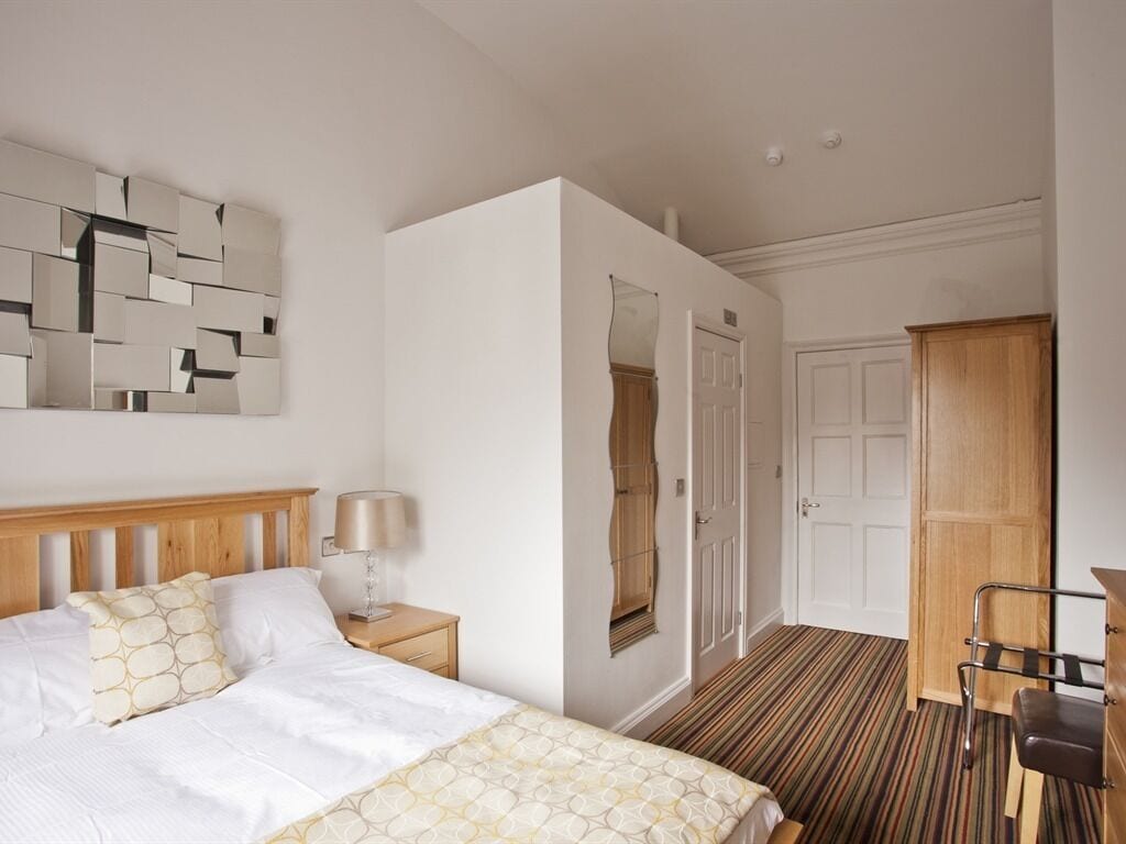 Een hotelkamer met een bed, nachtkastje, lamp, kledingkast en een spiegel aan de muur.