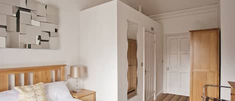 Een hotelkamer met een bed, nachtkastje, lamp, kledingkast en een spiegel aan de muur.