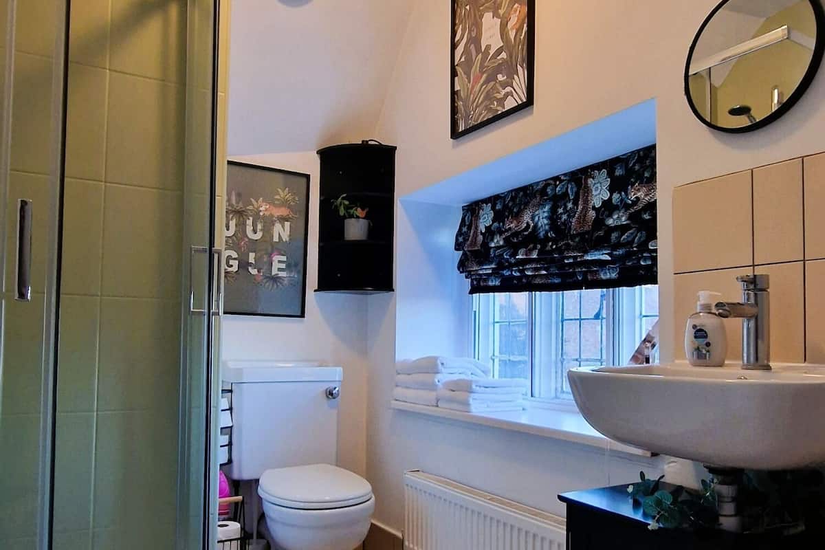 Ett badrum med en duschkabin i glas, en toalett, ett handfat med en stor spegel och ett fönster med en dekorativ gardinkappa.
