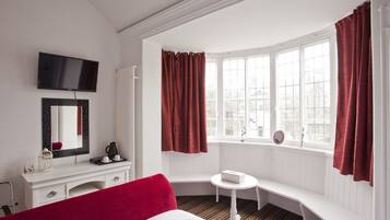 A hotel room with a bed, a white desk with a mirror, a television, a window with curtains, and a bench by the window.