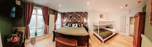 A bedroom with a large bed, a smaller bed on a platform, a window with curtains, a radiator, and a wall with a floral wallpaper.