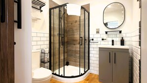 Un baño moderno con ducha envuelta en vidrio, azulejos blancos, suelo de madera, inodoro blanco y tocador con espejo e iluminación.