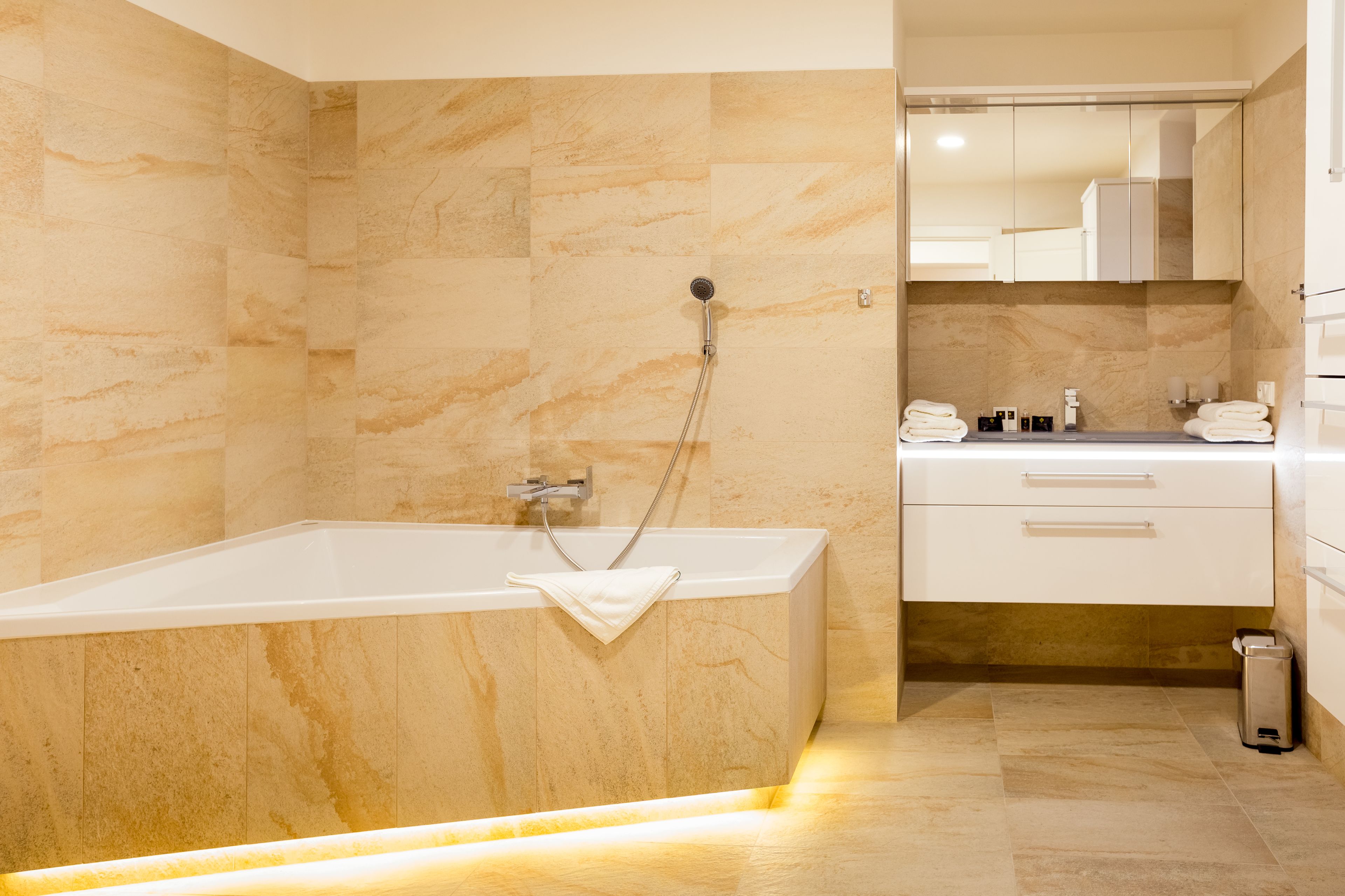 A modern bathroom with a marble wall, a white bathtub, a vanity with a mirror, and a trash can.