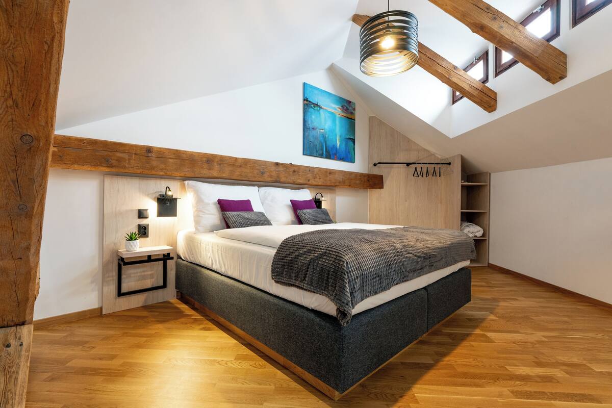 A modern loft bedroom with a sloped ceiling, exposed wooden beams, a large bed with a gray and white bedding, a small bedside table with a lamp, and a skylight above the bed.