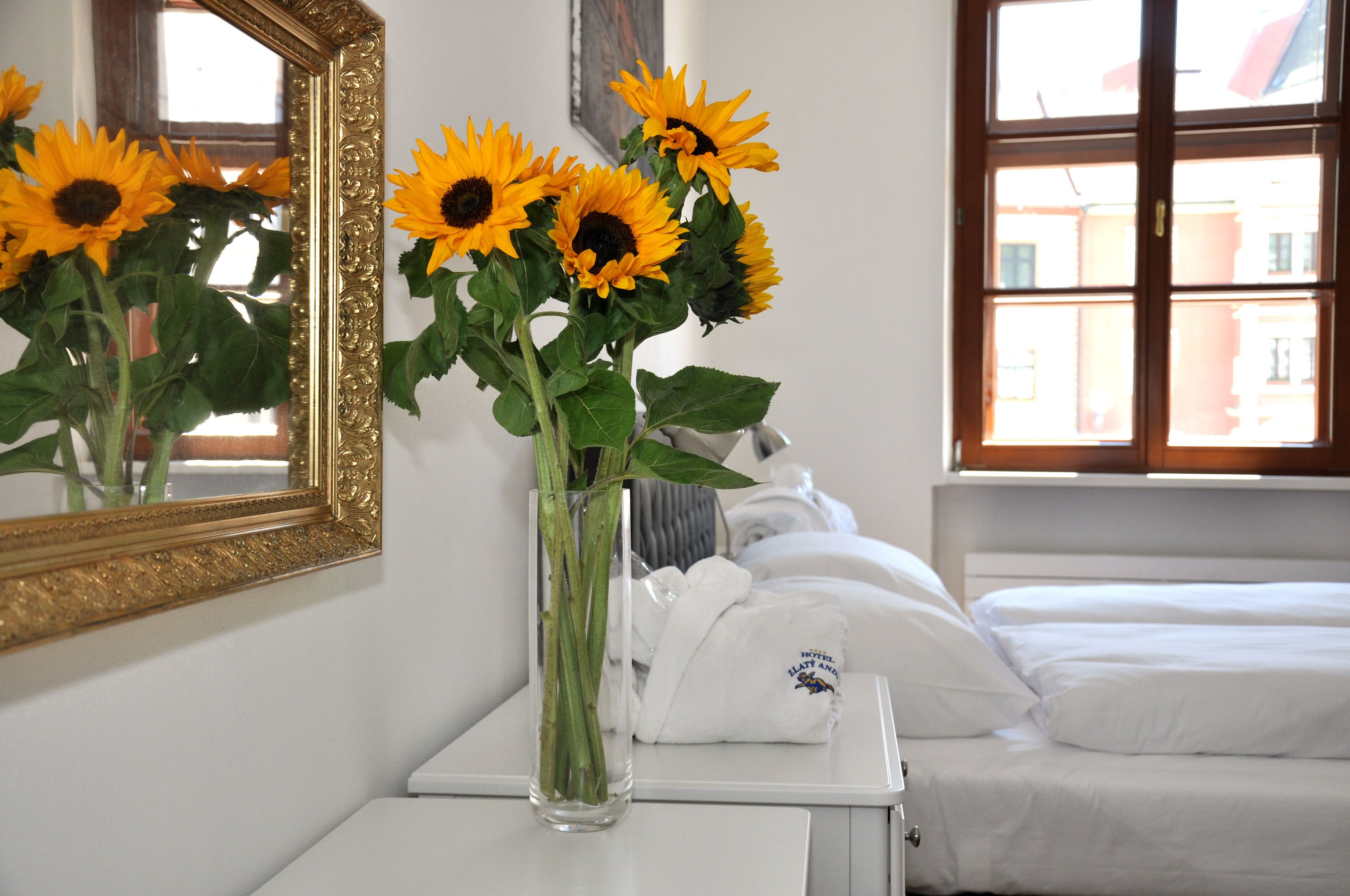 Habitación de hotel con cama, cómoda blanca, un jarrón con girasoles y un espejo con marco decorativo.
