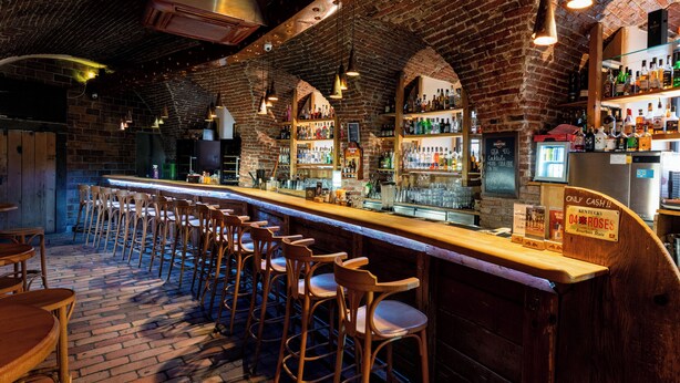 A bar with wooden stools, a brick wall, and shelves stocked with bottles. - Hotel Zlaty Andel (Cesky Krumlov)
