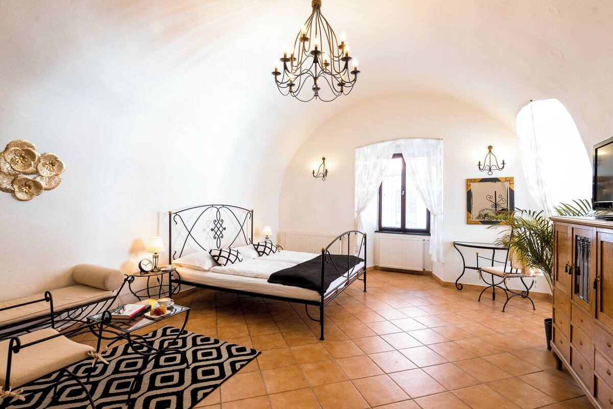 A compact room with a bed, a sofa, a small table with a lamp, a chandelier, and a cabinet.