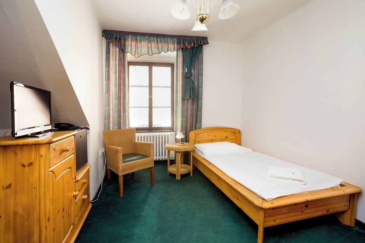 A small, tidy room with a single bed, a chair, a small table, and a wooden dresser.