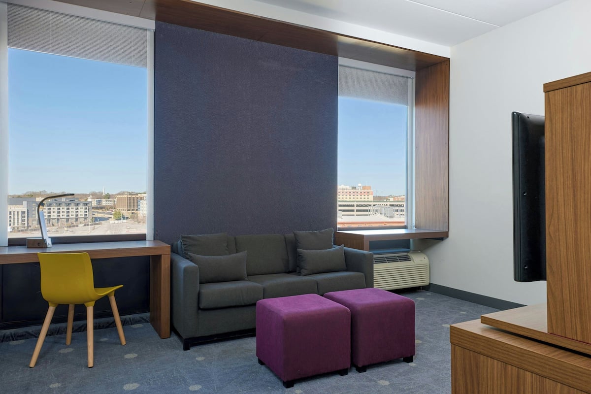 A modern hotel room with a large window, a sofa, a chair, a TV, and a small table.
