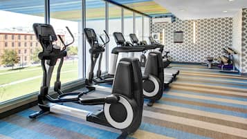 Un gimnasio con cintas de correr, una alfombra estampada y amplios ventanales con vistas a un edificio y una calle.