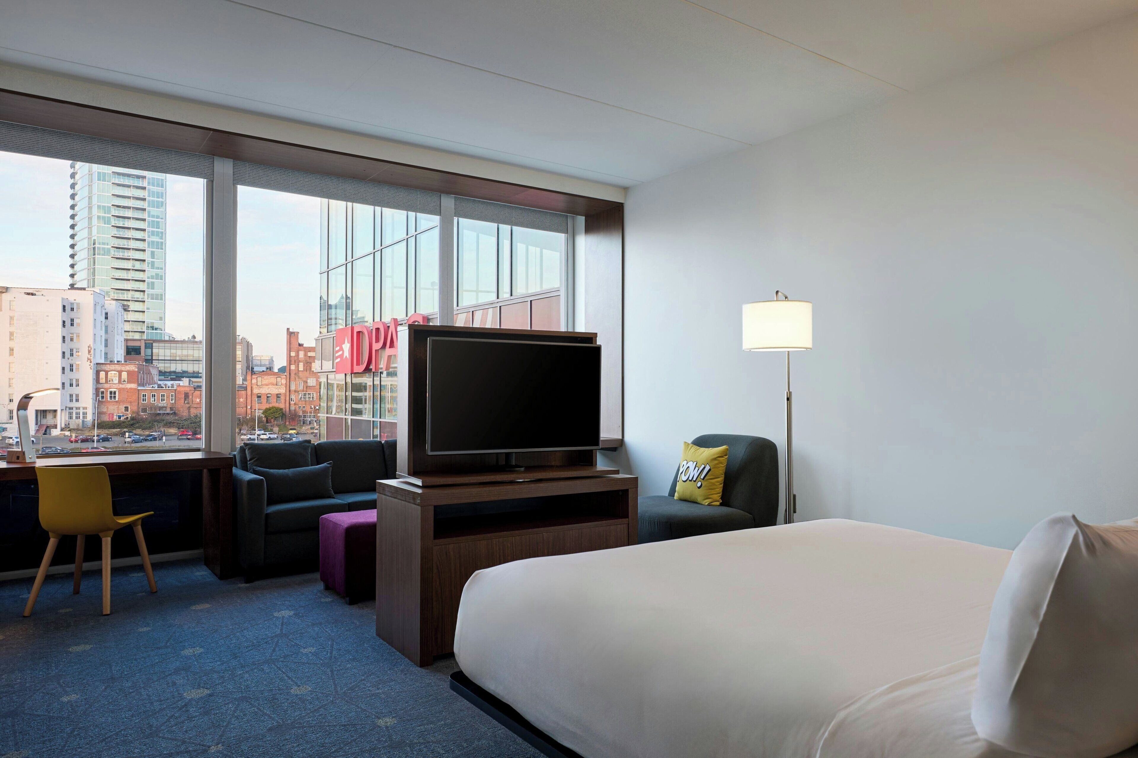Quarto de hotel com uma cama grande, televisão, escrivaninha com cadeira, sofá e janela com vista para a cidade.