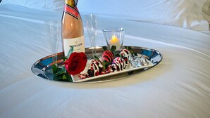 Ein ordentlich bezogenes Bett mit weißen Leinentüchern, eine Champagnerflasche, ein Glas sowie ein Tablett mit Erdbeeren und einer Kerze in einem Hotelzimmer.