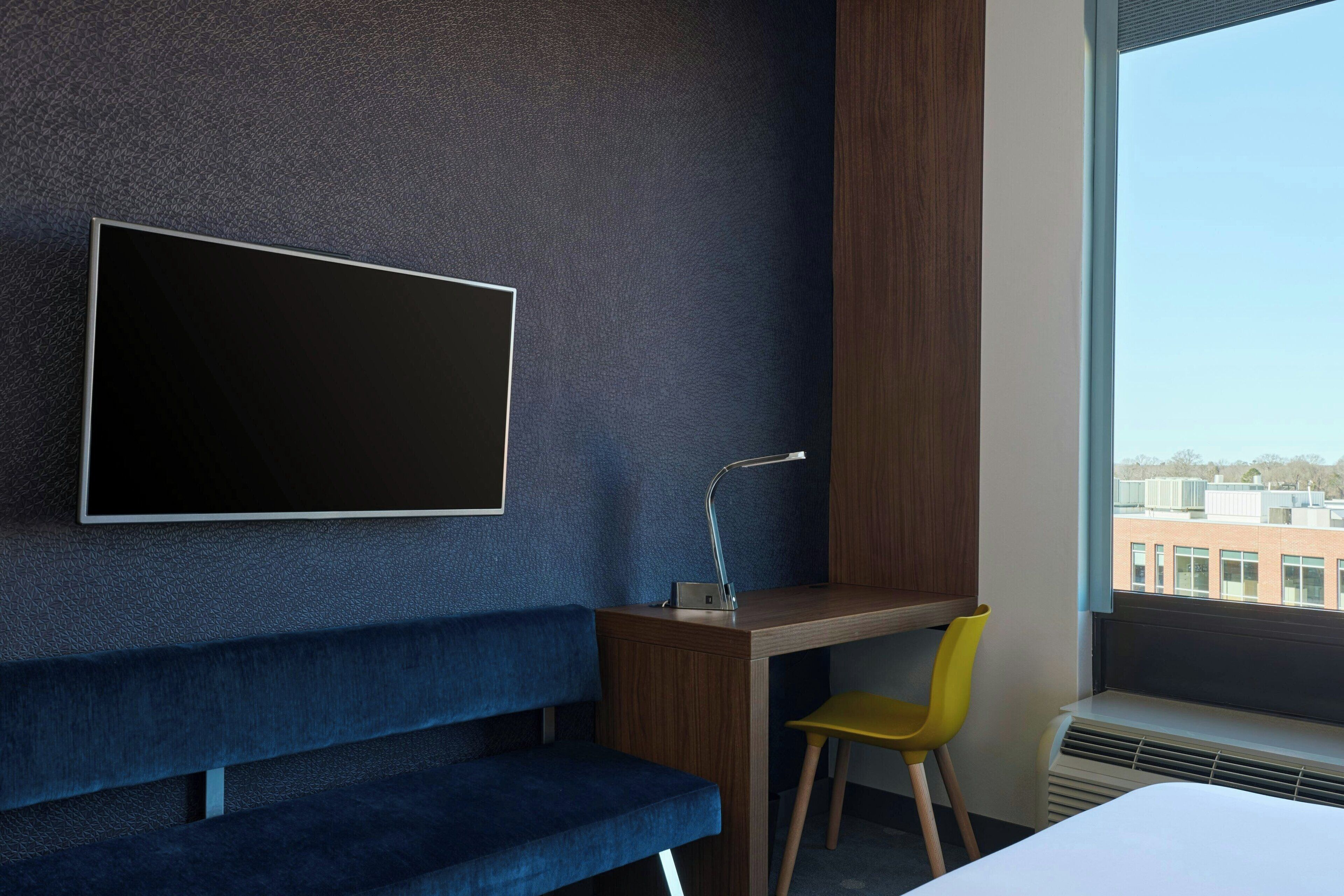 Quarto de hotel moderno com uma televisão grande fixada na parede, uma escrivaninha de madeira, um banco azul, uma cadeira amarela e uma janela com vista para a cidade.