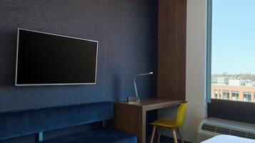 Quarto de hotel moderno com uma televisão grande fixada na parede, uma escrivaninha de madeira, um banco azul, uma cadeira amarela e uma janela com vista para a cidade.