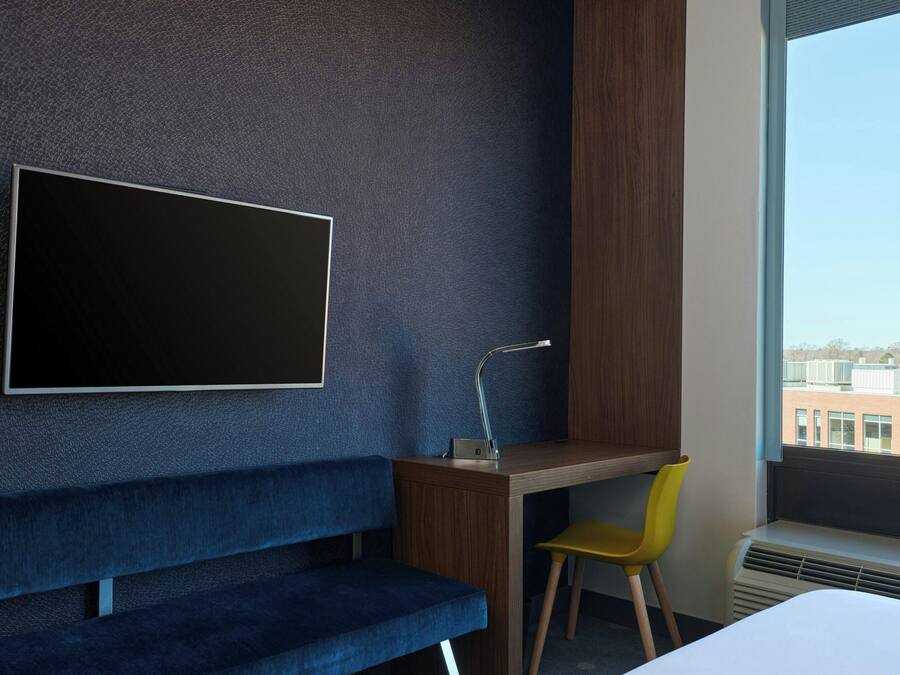 Quarto de hotel moderno com uma televisão grande fixada na parede, uma escrivaninha de madeira, um banco azul, uma cadeira amarela e uma janela com vista para a cidade.