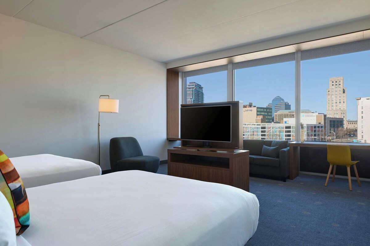 Habitación de hotel con dos camas, televisión, escritorio y ventana con vista a la ciudad.