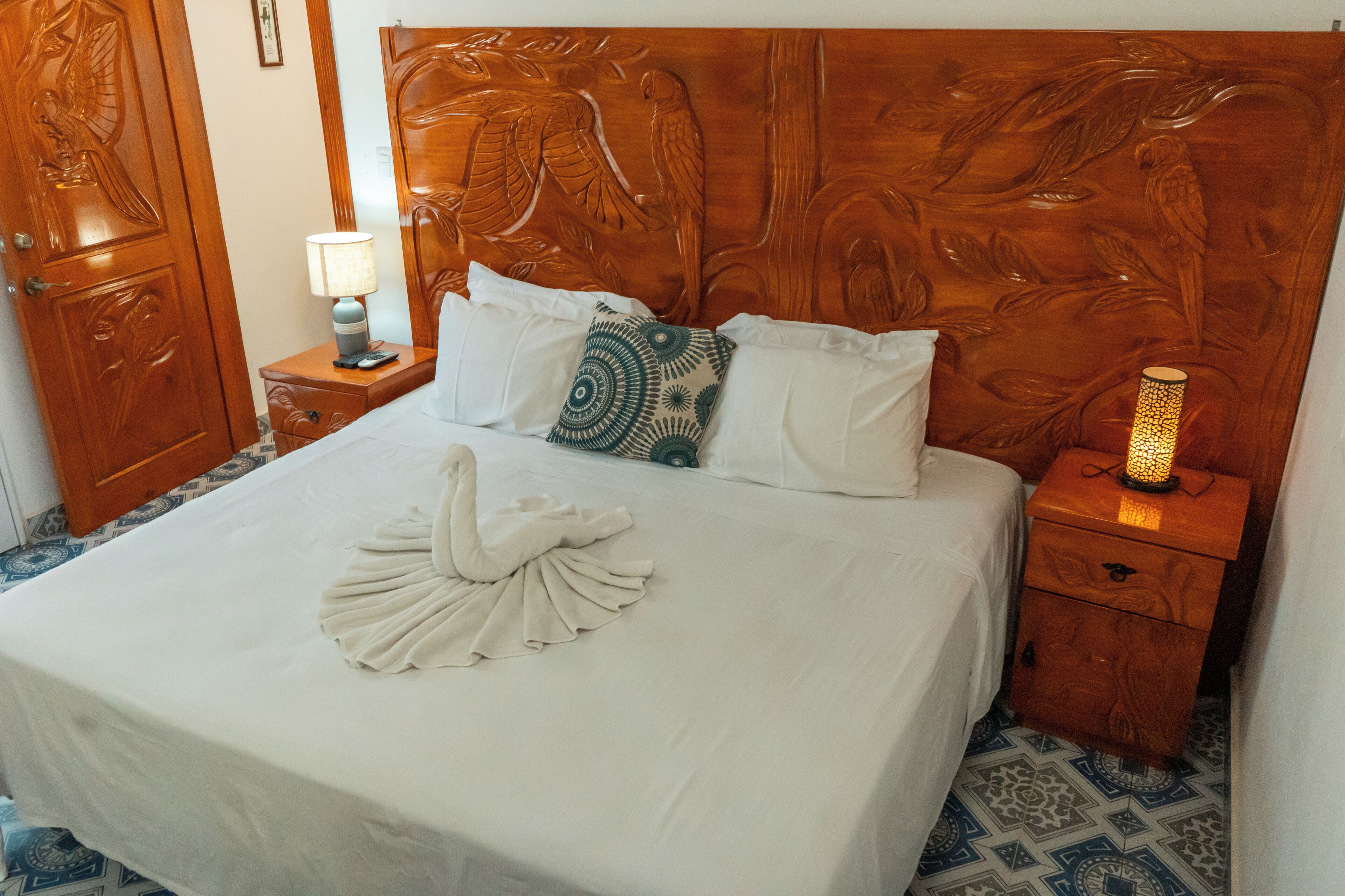 A hotel room with a wooden headboard, a bed with white bedding, a decorative swan pillow, a nightstand with a lamp, and a patterned floor.