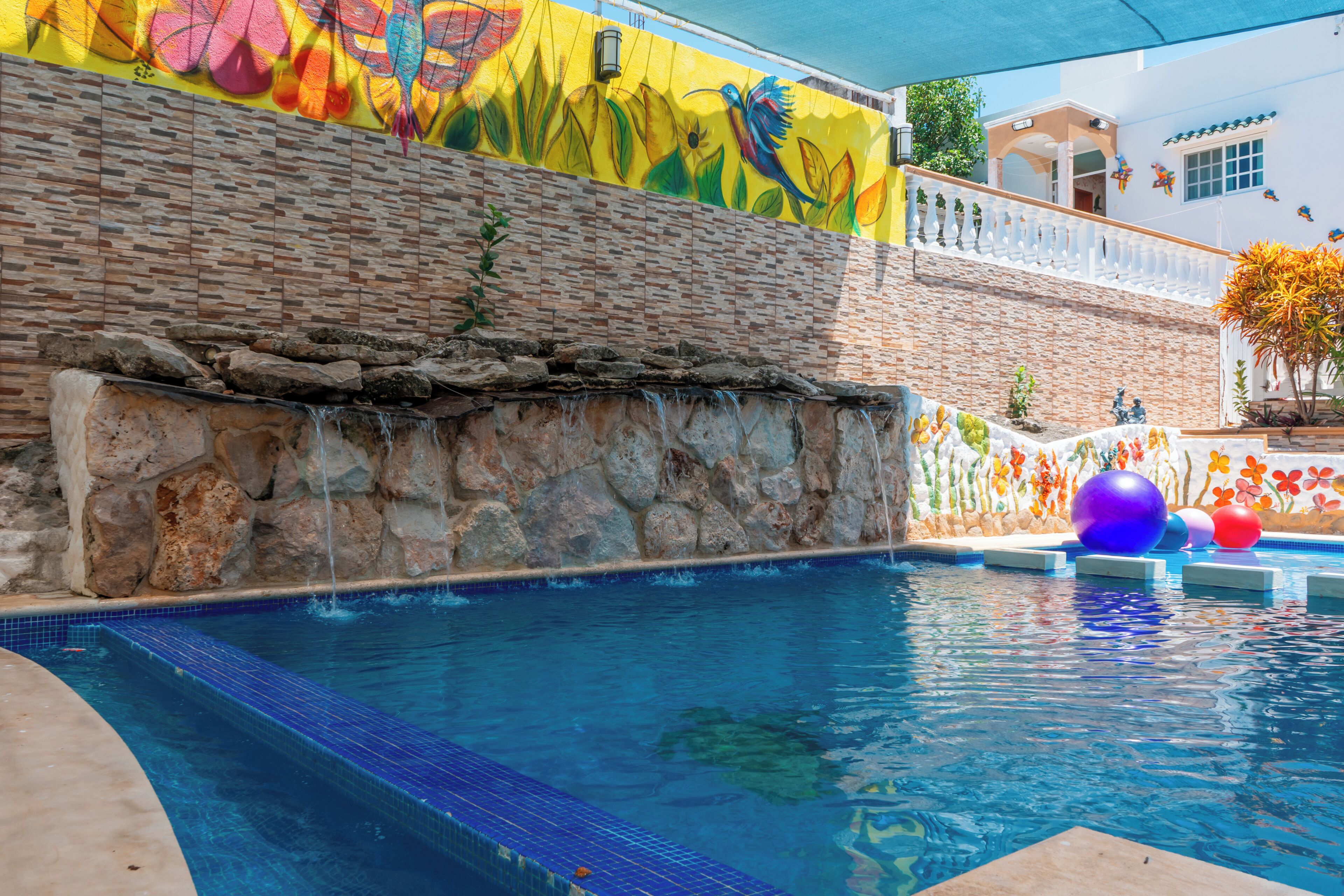 A swimming pool with a stone wall and colorful mural, surrounded by a white fence and a building with windows.