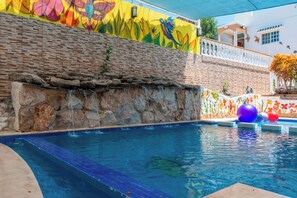 Une piscine bordée d’un mur en pierre et agrémentée d’une fresque colorée, entourée d’une clôture blanche et d’un bâtiment doté de fenêtres.