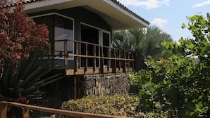 A house with a balcony, stone foundation, and surrounding tropical vegetation.