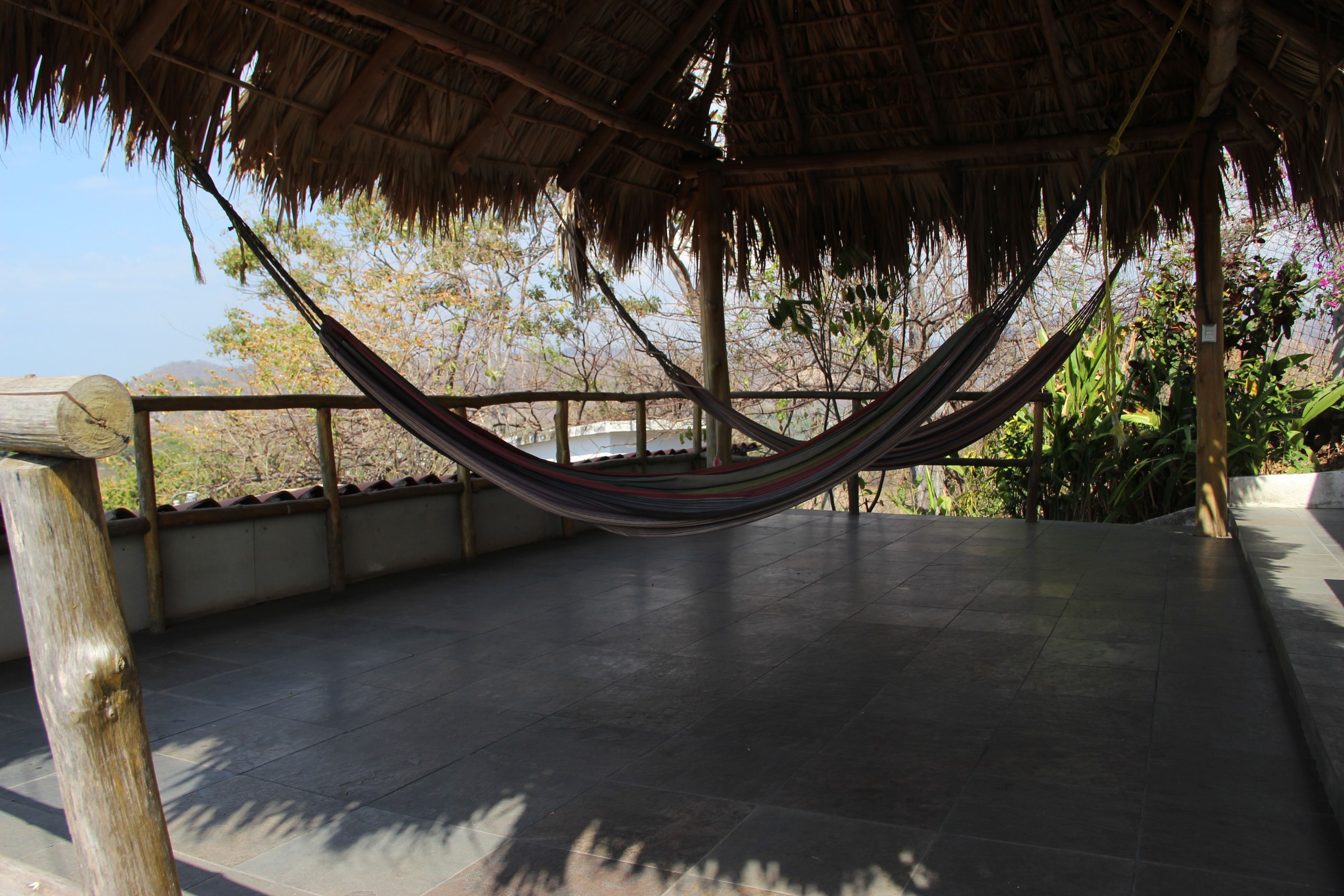 A hammock under a thatched roof structure, with a tiled floor and a view of trees and hills in the background.