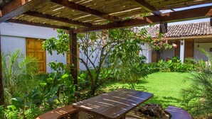 Un porche cubierto con una mesa y bancos de madera, rodeado de exuberante vegetación y un edificio blanco con techo de tejas.