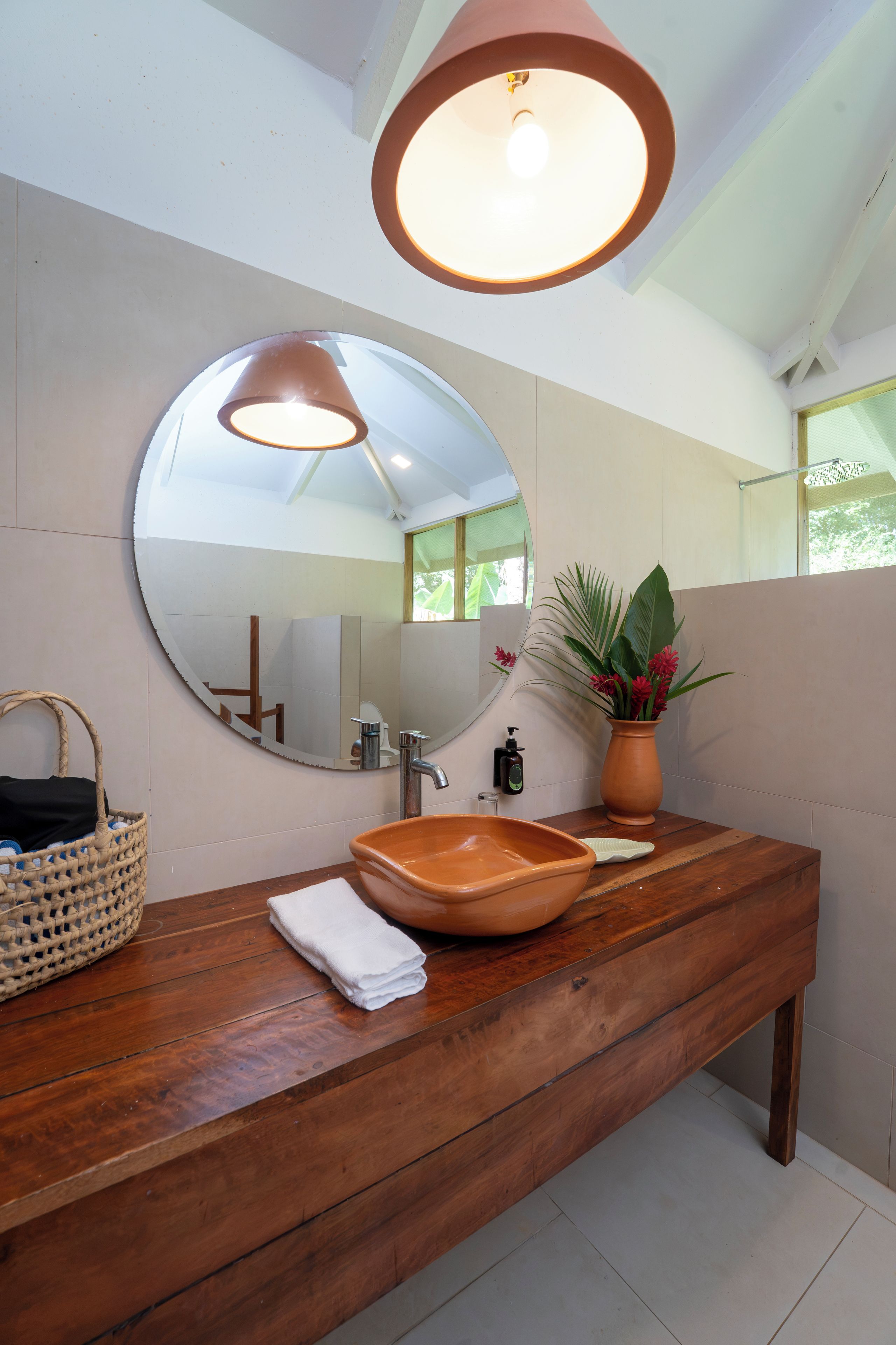 A bathroom with a wooden vanity, a round mirror, a towel, a soap dispenser, and a potted plant.