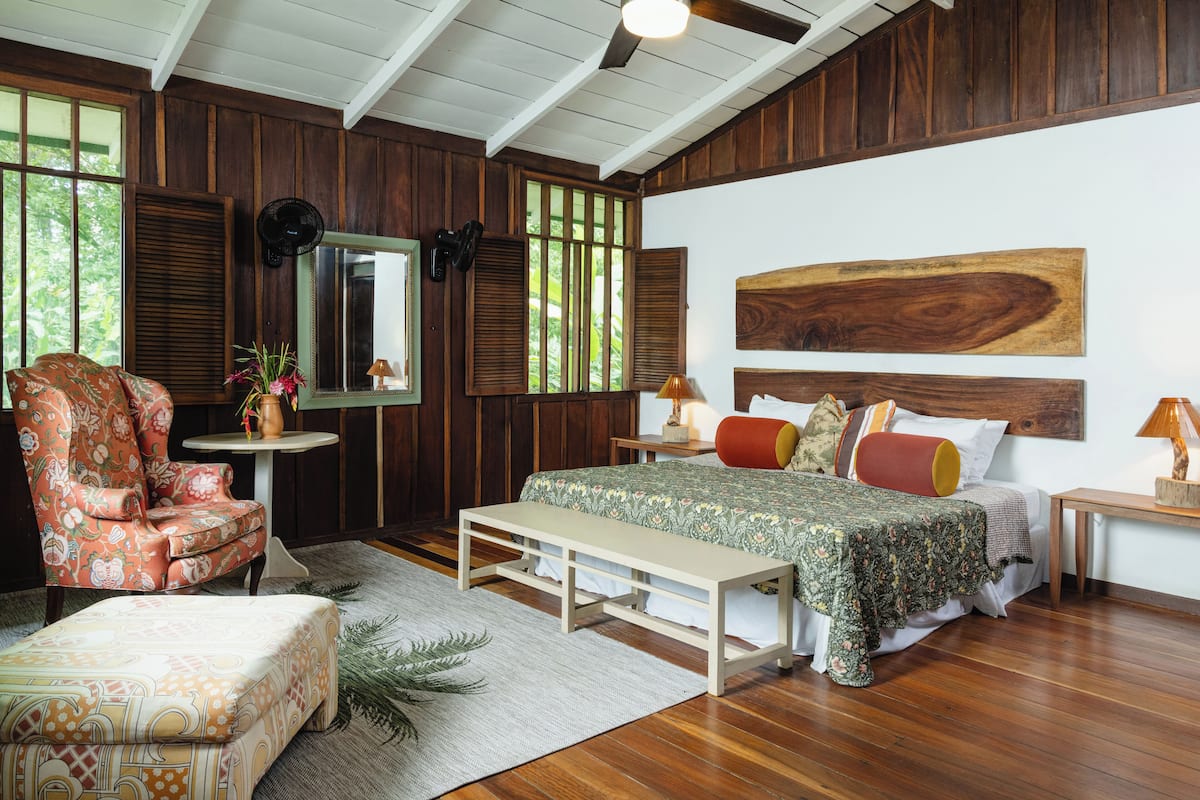 Ein Schlafzimmer mit Holzbalkendecke, einem großen Bett mit floralem Bettzeug, einer Bank, einem Stuhl und einem Spiegel an der Wand.