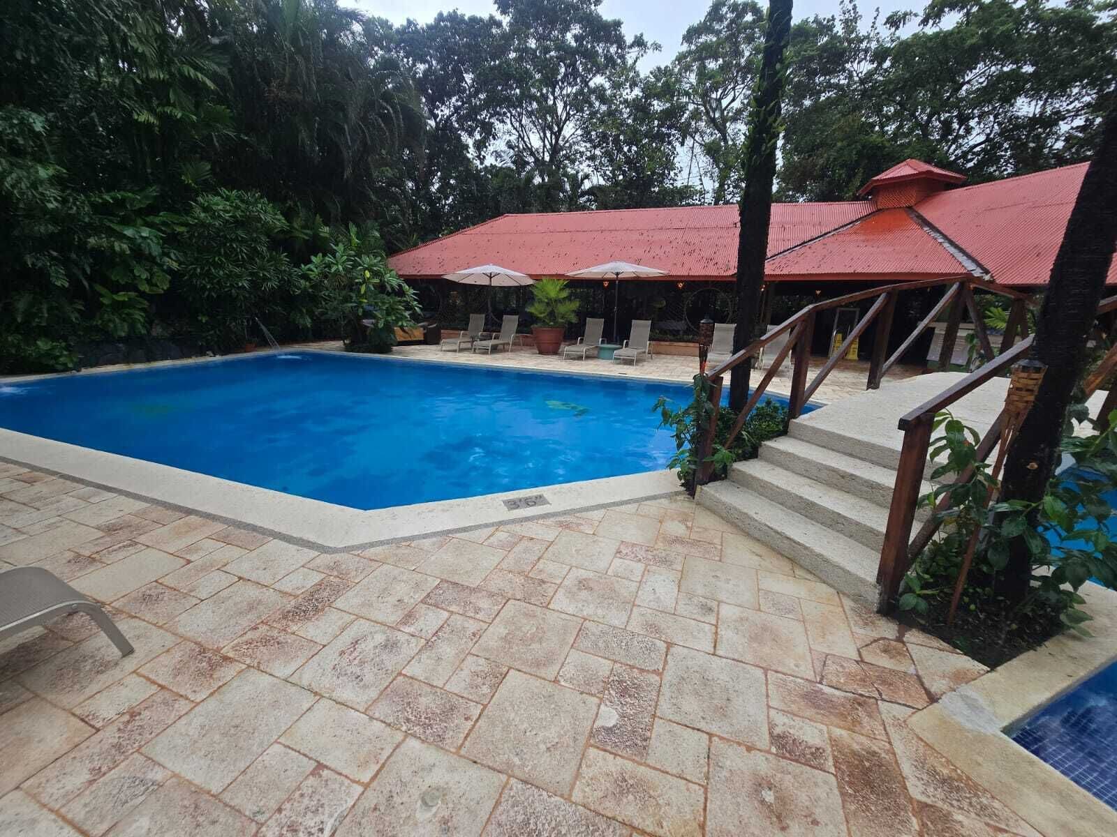 Una piscina con una estructura de techo rojo, suelo de piedra y una escalera que lleva a una zona cubierta.