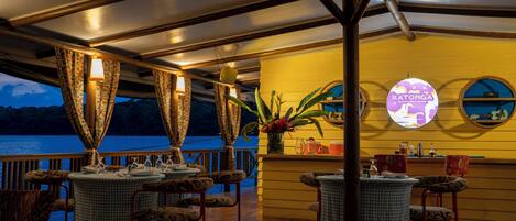 A restaurant with tables and chairs, a view of the sea, and a sign that reads 'Katonga.'